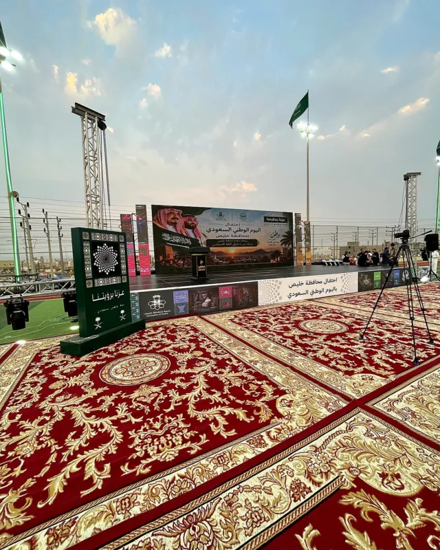 ساحة احتفال اليوم الوطني في خليص مفروشة بسجاد أحمر ملكي، مع المسرح والشاشة وأعلام المملكة في الخلفية