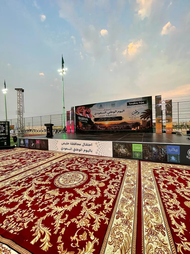 مسرح احتفال محافظة خليص باليوم الوطني السعودي بشاشة كبيرة، وأمامه ساحة مفروشة بسجاد أحمر ملكي بنقوش ذهبية