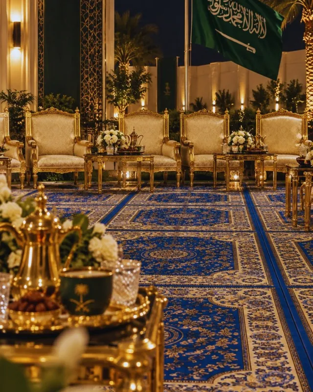 جلسة استقبال ليلية بكنب ملكي أبيض بإطار ذهبي على سجاد أزرق ملكي بنقوش ذهبية، مع دلال قهوة ذهبية وعلم المملكة في الخلفية