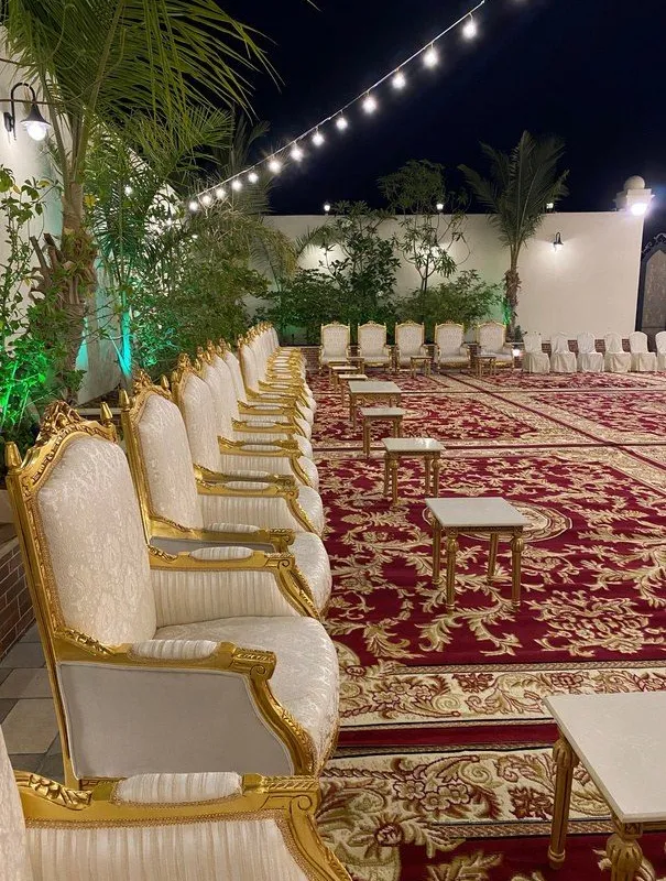 مجلس خارجي مسائي بكنب ملكي أبيض مذهّب مصفوف على سجاد أحمر بنقوش ذهبية، مع طاولات خدمة رخامية وعقود إنارة فوق الجلسات