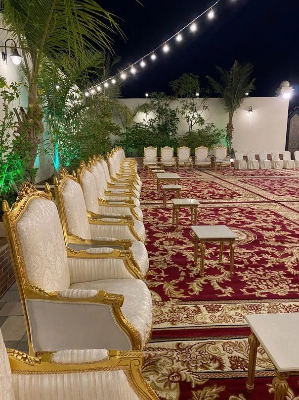 مجلس خارجي مسائي بكنب ملكي أبيض مذهّب مصفوف على سجاد أحمر بنقوش ذهبية، مع طاولات خدمة رخامية وعقود إنارة فوق الجلسات