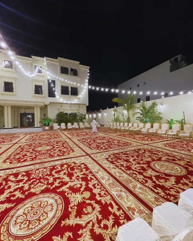 فناء فيلا مجهز لمناسبة ليلية بسجاد أحمر ملكي يغطي الساحة، وصفوف كراسي مغطاة بيضاء، وعقود إنارة ممتدة فوق المكان