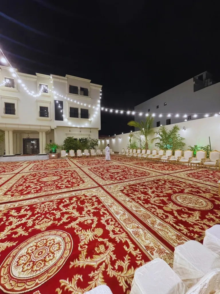 فناء فيلا مجهز لمناسبة ليلية بسجاد أحمر ملكي يغطي الساحة، وصفوف كراسي مغطاة بيضاء، وعقود إنارة ممتدة فوق المكان