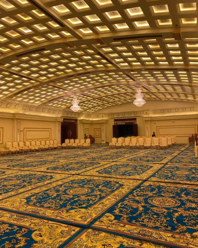 قاعة مناسبات واسعة مفروشة بالكامل بسجاد أزرق ملكي بنقوش ذهبية، مع صفوف جلسات في الخلف وسقف بإضاءة دافئة