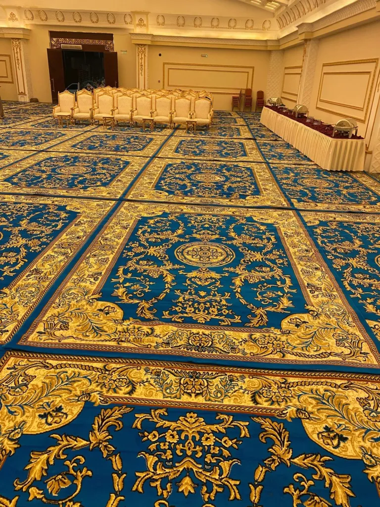 سجاد أزرق ملكي بزخارف ذهبية مفروش في قاعة مناسبات، وفي الخلفية كراسٍ مصفوفة وطاولة ضيافة